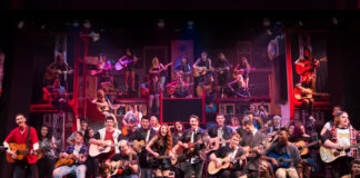A large group of people of various ages and genders play acoustic guitars on a colorful, vibrant stage set, some seated and some standing, with shelves and props in the background.