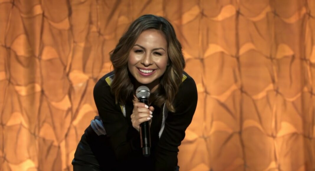 A woman with wavy brown hair smiles and leans forward while holding a microphone on stage, with a patterned golden curtain as the background.