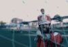 A drum major in a red and white uniform and tall hat points a baton toward the camera through a soccer net on an outdoor field, with blurred structures and people in the background.