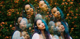 A woman with long blue and purple hair stands in front of colorful flowers, her image artistically duplicated and layered multiple times to create a surreal, kaleidoscopic effect.