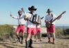 Four men wear white shirts, pink shorts, and boots while playing musical instruments outdoors. One plays an accordion, another a bass guitar, a third plays drums, and the fourth holds drumsticks, with cliffs and blue sky in the background.