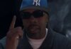A man in a blue New York Yankees cap and dark sunglasses raises his index finger while looking at the camera, with smoke swirling in the background.