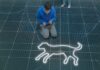 A person in a blue hoodie kneels on a grid-patterned floor, looking at a glowing outline of a dog drawn on the ground. Another persons legs are visible standing nearby.