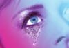 A close-up of a person’s face with bright blue eyes, sparkling eyeshadow, and glittery tears streaming down their cheek. The image has a vibrant pink and blue color overlay.