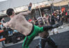 A wrestler in green pants delivers a high kick to an opponent wearing a colorful mask during an outdoor match, as spectators watch from behind the ring ropes.