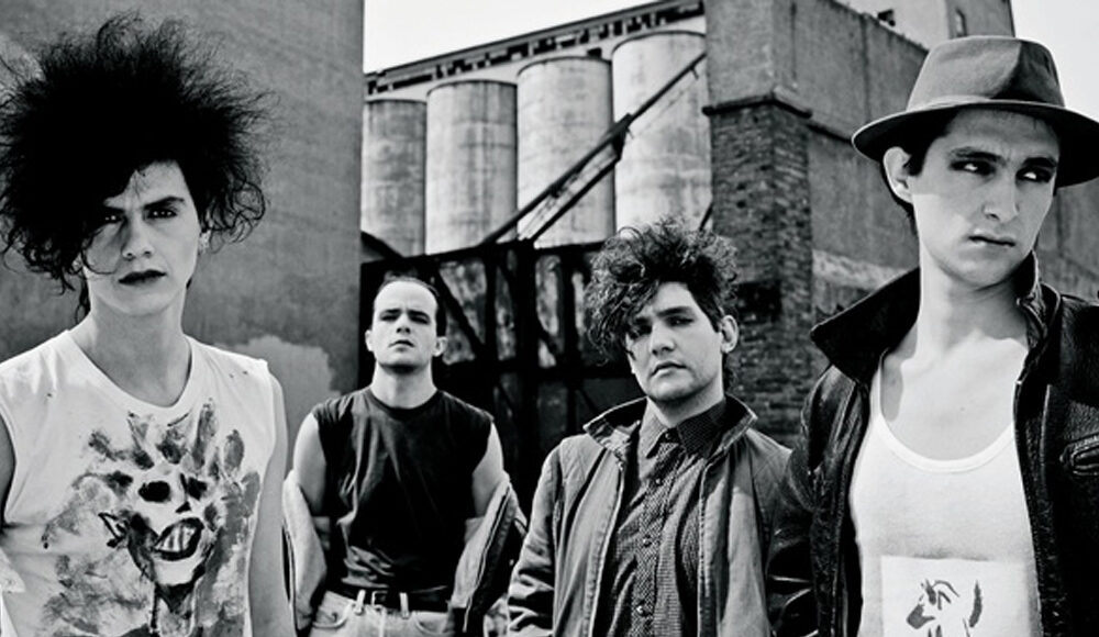 Four young men with 1980s punk and new wave fashion stand outside in an industrial area. One has big, teased hair and a graphic t-shirt, another wears a sleeveless shirt, and one wears a jacket and another a hat.