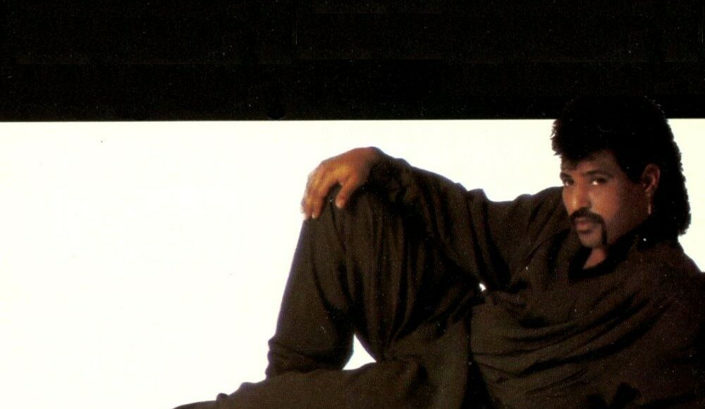 A man with a mustache and mullet hairstyle lounges on the floor in a relaxed pose, wearing a loose-fitting dark outfit and white shoes against a plain white background.