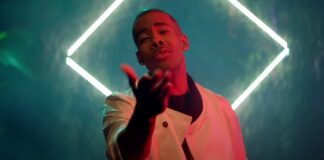 A man in a white jacket reaches his hand toward the camera, standing in front of a glowing diamond-shaped light with a misty, colorful background.