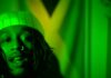 A person with a nose ring and a beanie is shown in green lighting, with a Jamaican flag in the background.