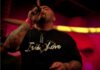 A person with tattoos sings into a microphone on stage, wearing a black shirt that says Irie Love in white letters, with a necklace and a red wristband. The background is dimly lit in warm, reddish tones.
