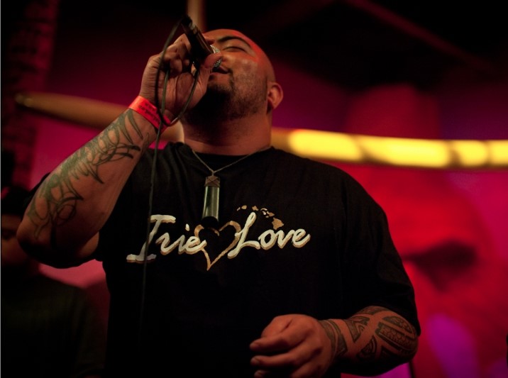 A person with tattoos sings into a microphone on stage, wearing a black shirt that says Irie Love in white letters, with a necklace and a red wristband. The background is dimly lit in warm, reddish tones.