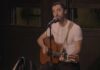A man wearing a white t-shirt plays an acoustic guitar and sings into a microphone on stage. A jacket hangs on the back of his chair, and he appears focused on his performance.