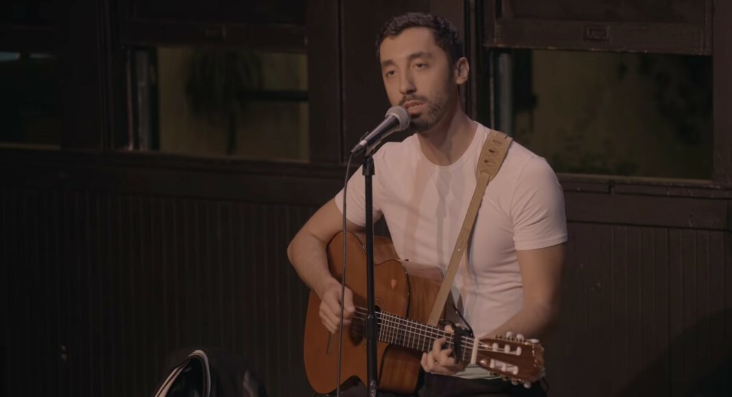 A man wearing a white t-shirt plays an acoustic guitar and sings into a microphone on stage. A jacket hangs on the back of his chair, and he appears focused on his performance.