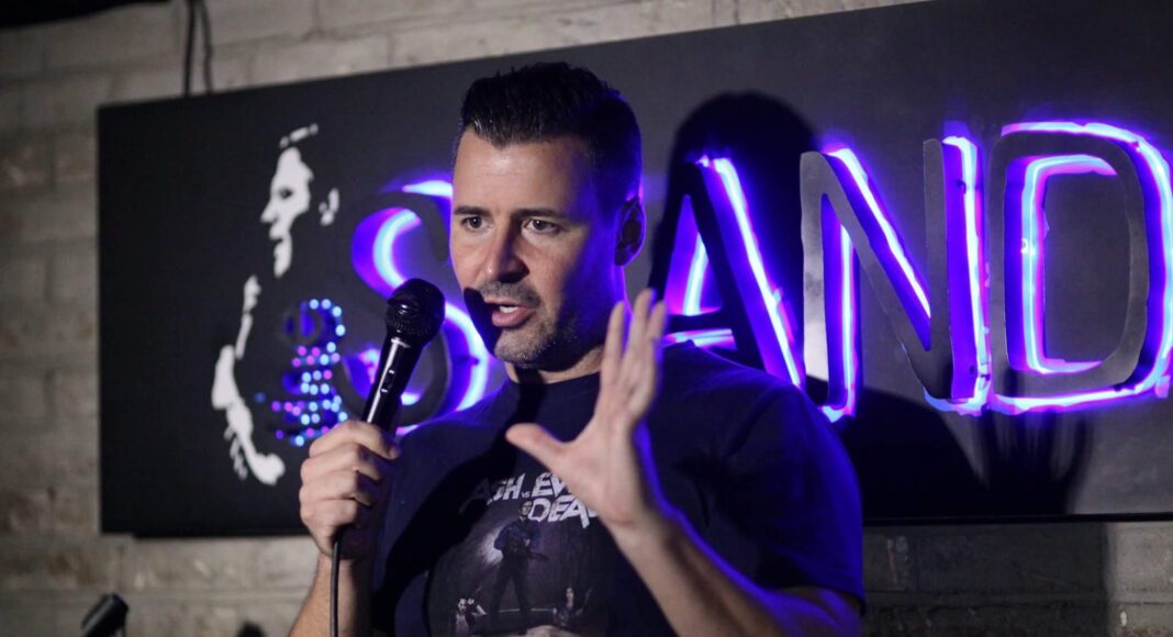 A man holding a microphone performs stand-up comedy on stage, with a neon STAND sign glowing in blue behind him and a brick wall in the background.