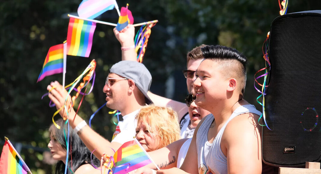 A diverse group of people smile and wave rainbow pride flags at an outdoor event, with colorful streamers and a large speaker visible nearby.