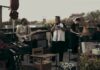 A band of five musicians performs on a rooftop. One plays keyboard, another guitar, one trumpet, one drums, and another handles electronic equipment. Plants and chairs decorate the space, with a city skyline in the background.