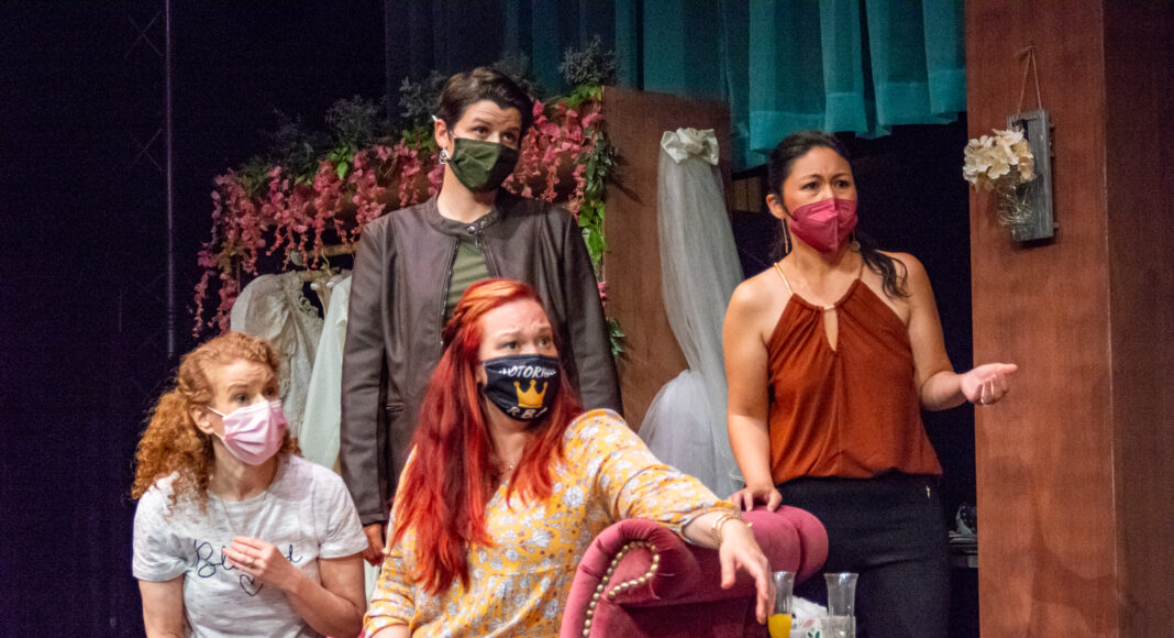 Four women wearing face masks are gathered in a room decorated with flowers and fabric. Three stand, and one with red hair sits on a pink couch. They appear to be engaged in conversation or reacting to something.