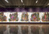 A large, colorful mixed-media art installation featuring five panels filled with vibrant, dense clusters of artificial flowers and butterflies, set against a purple, floral-patterned wall in a spacious gallery.