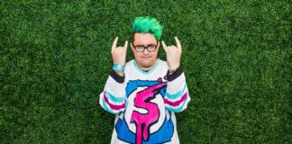 Person with bright green curly hair and glasses stands in front of a leafy green background, wearing a colorful graphic jersey and making rock and roll hand signs with both hands.