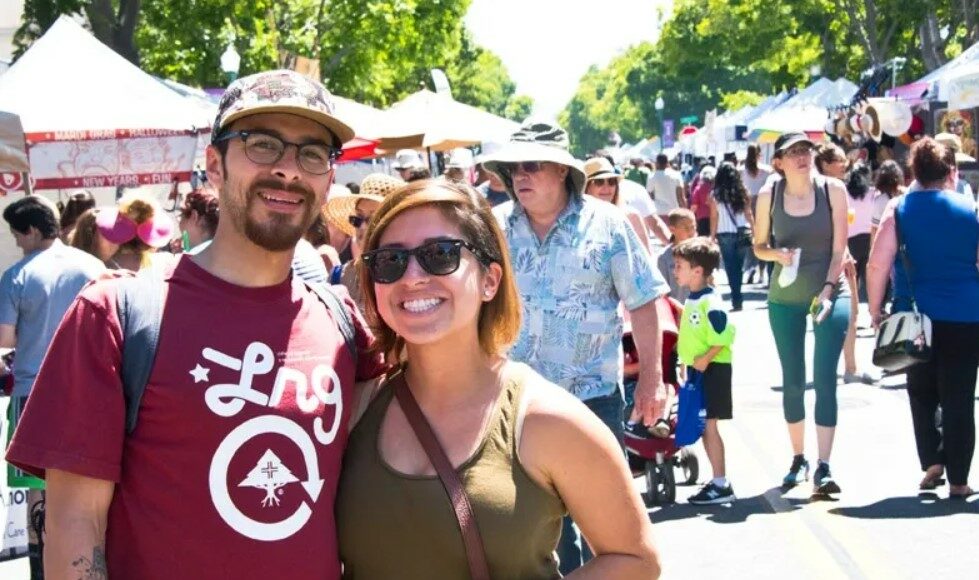 A smiling man and woman pose together at an outdoor street fair, surrounded by people, tents, and booths on a sunny day. The man wears glasses and a red shirt, and the woman wears sunglasses and a green tank top.