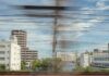 View of modern city buildings and power lines seen through a train window, with motion blur suggesting the train is moving quickly. Blue sky with some clouds is visible in the background.