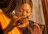 A woman wearing a microphone headset plays a harp, her fingers plucking the strings as she focuses intently on the instrument. The harps strings and frame are visible in the foreground.