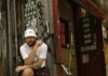 A man in a white bucket hat and casual clothes sits on a step next to a bike, in front of a worn building with peeling posters, Chinese text, and phone numbers on the walls.