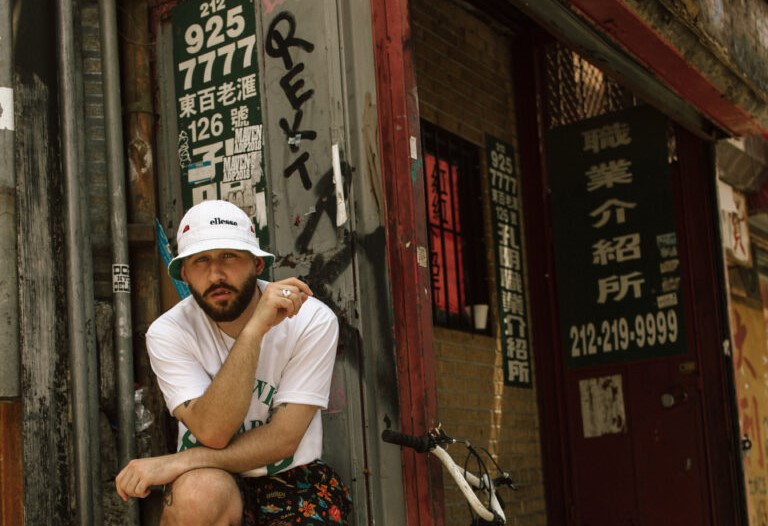 A man in a white bucket hat and casual clothes sits on a step next to a bike, in front of a worn building with peeling posters, Chinese text, and phone numbers on the walls.