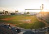 A baseball game takes place in a stadium at sunset. Players are spread across the field, and a small crowd watches from the stands. The sky is clear, and sunlight illuminates the field and empty seats.