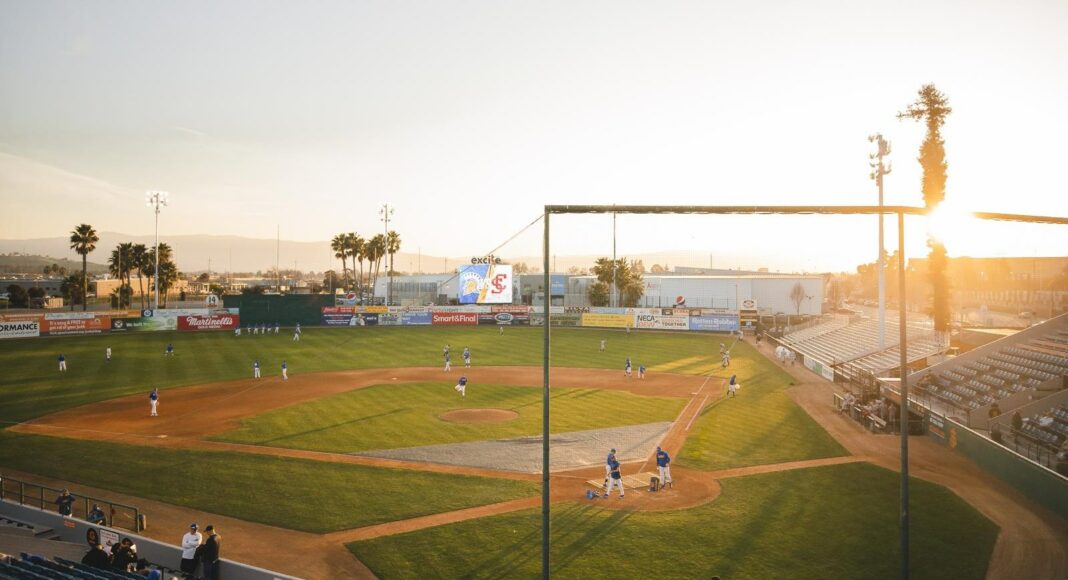 A baseball game takes place in a stadium at sunset. Players are spread across the field, and a small crowd watches from the stands. The sky is clear, and sunlight illuminates the field and empty seats.