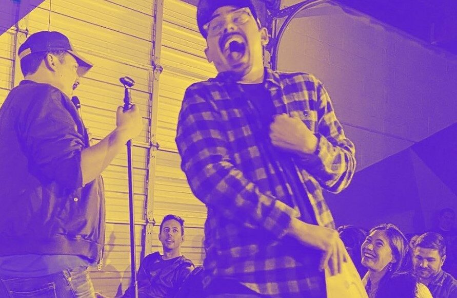 A man with a microphone stands on stage as another man in a plaid shirt reacts dramatically, mouth open wide. Several people sitting nearby laugh and watch the scene unfold. The image has a yellow and purple filter.