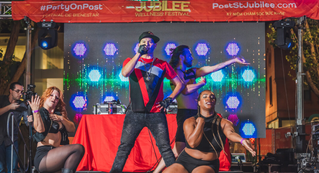 A performer in a red and black outfit sings on an outdoor stage with a DJ behind him, while two dancers in black pose energetically at the front. Bright lights and festival banners are visible in the background.