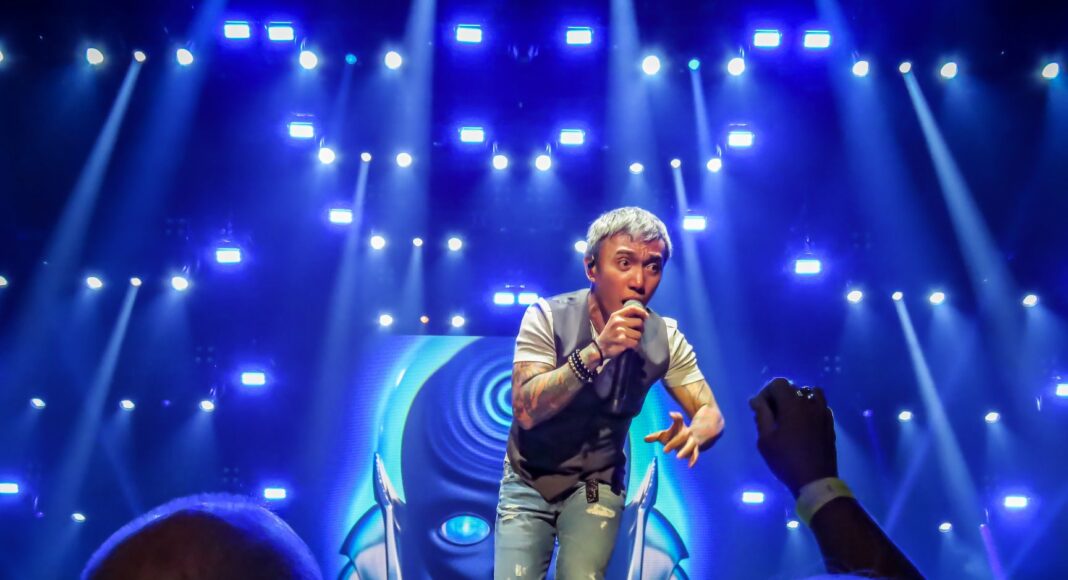 A singer with short, light-colored hair performs energetically on stage under bright blue lights, while fans in the audience watch and take photos. A futuristic graphic is displayed on a large screen behind him.