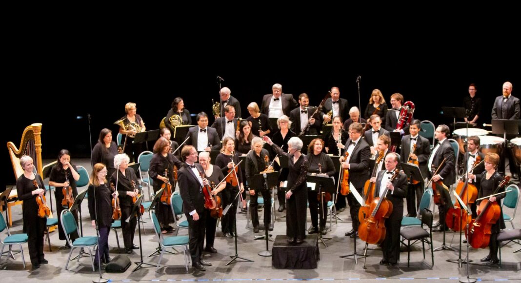A full orchestra stands on stage with their instruments, wearing formal black attire. The conductor stands at the center front, and the musicians are arranged in sections, smiling and acknowledging the audience.
