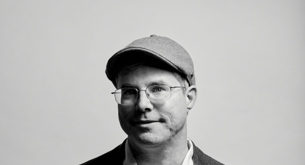 Black and white portrait of a man wearing glasses, a flat cap, a white shirt, and a dark blazer, facing the camera with a neutral expression against a plain background.