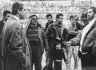 A group of men and a child stand outdoors in front of a graffiti-covered stone wall. One man holds a microphone toward another, suggesting an interview or news event. The people appear focused and engaged.