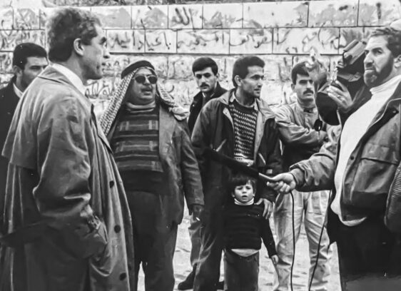 A group of men and a child stand outdoors in front of a graffiti-covered stone wall. One man holds a microphone toward another, suggesting an interview or news event. The people appear focused and engaged.