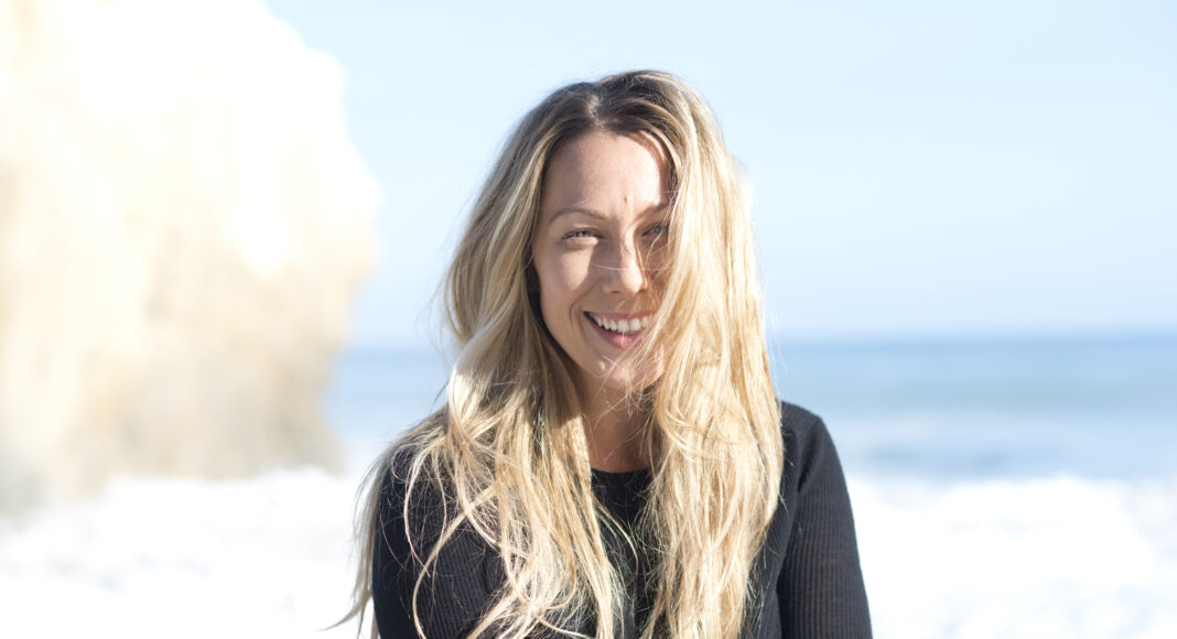 A woman with long blonde hair, wearing a black sweater, smiles at the camera while standing on a sunny beach with the ocean and a rock formation in the background.