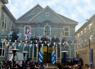 A festive courtyard with blue and white pennant banners and balloons, outdoor tables, and people gathered in front of a teal building decorated with various flags. String lights are hung across the open area.