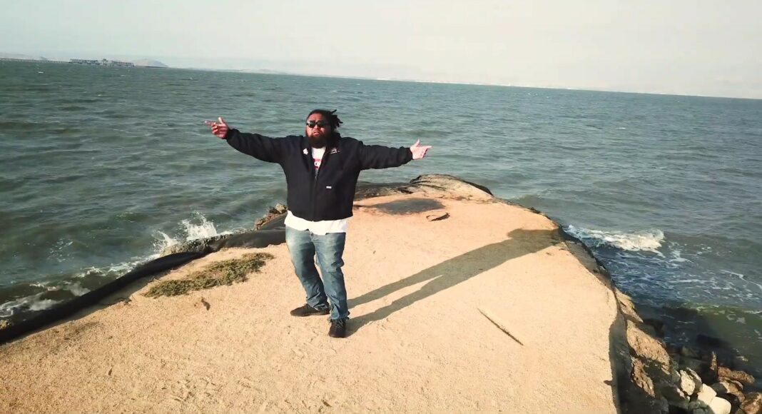 A person stands on a sandy, narrow pier extending into the ocean, arms outstretched. Waves crash on either side, and the sky is clear and bright. The figure casts a long shadow on the ground.
