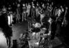 A drummer plays on stage while a large crowd stands close, watching intently. The scene is lit dramatically in black and white, highlighting the energy of the live performance and the audience’s engagement.