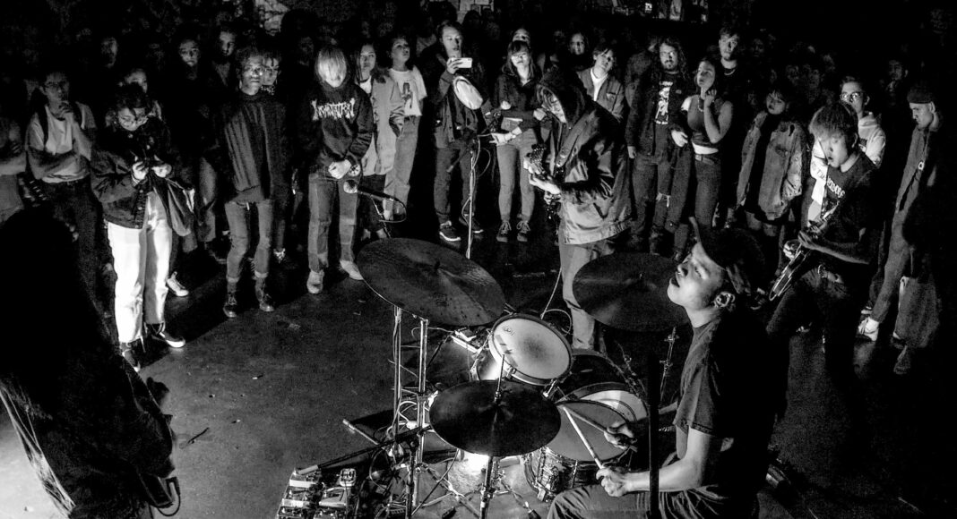 A drummer plays on stage while a large crowd stands close, watching intently. The scene is lit dramatically in black and white, highlighting the energy of the live performance and the audience’s engagement.