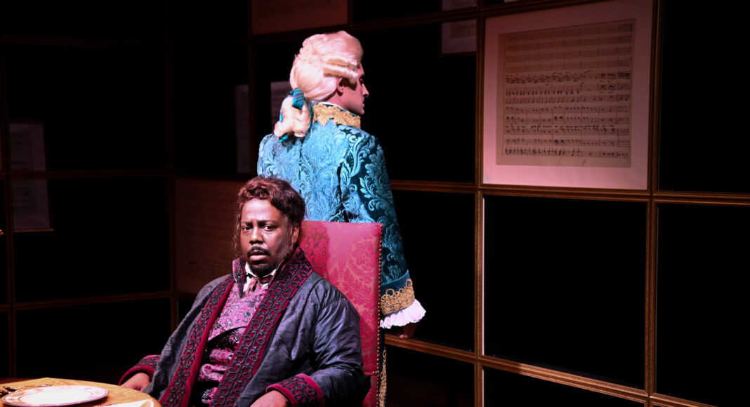 A man in an ornate robe sits in a red chair at a table, looking forward, while another person in an elaborate blue 18th-century costume with a white wig stands with their back turned, in a room with framed sheet music on the walls.