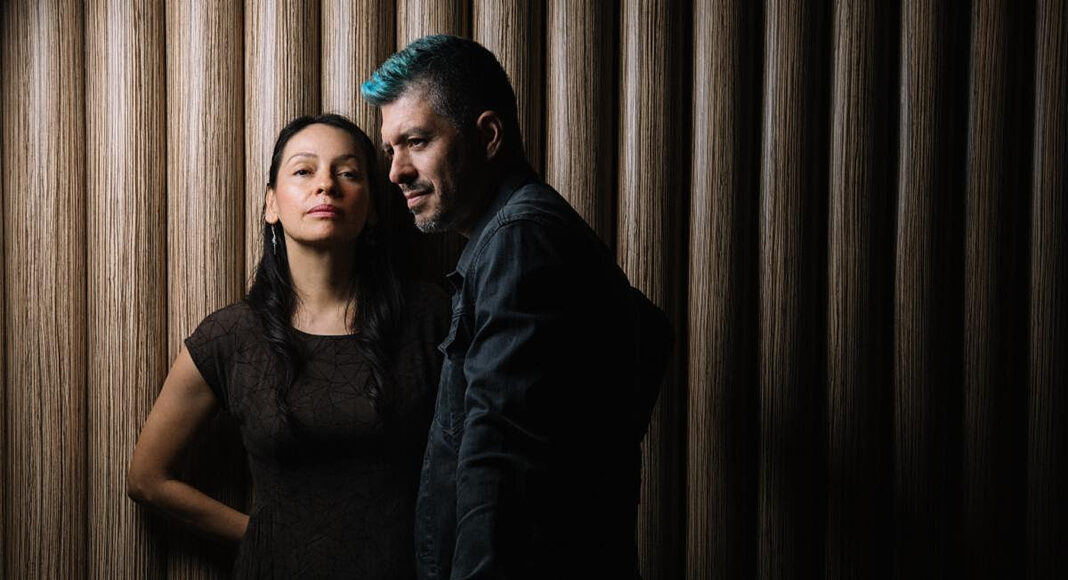 A woman in a dark dress stands next to a man with blue hair in a dark jacket. They pose against a background of vertical wooden panels, both looking forward with serious expressions.