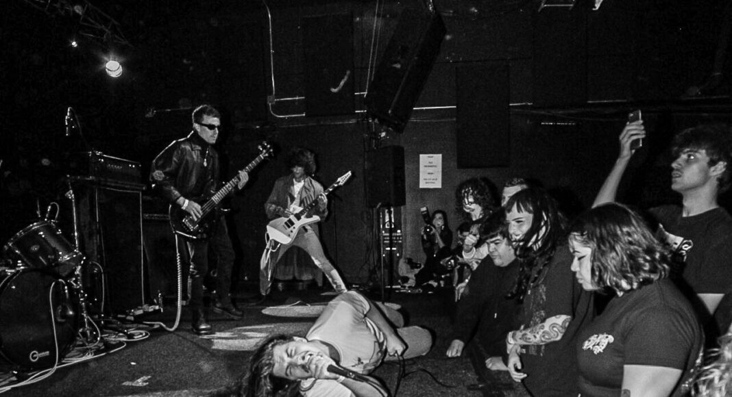 A black and white photo of a band performing on stage. The vocalist lies on the floor while singing, with enthusiastic audience members reaching toward the stage. Two guitarists play in the background.