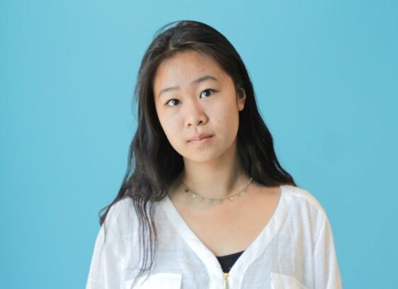 A young woman with long dark hair wearing a white shirt and a delicate necklace stands in front of a solid light blue background, looking slightly to the side with a neutral expression.