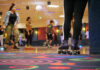 People roller skating in an indoor rink with colorful carpet and a disco ball overhead. In the foreground, a person wearing black leggings and skates stands near the edge, watching others skate.