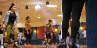 People roller skating in an indoor rink with colorful carpet and a disco ball overhead. In the foreground, a person wearing black leggings and skates stands near the edge, watching others skate.