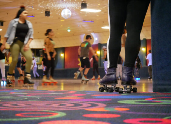 People roller skating in an indoor rink with colorful carpet and a disco ball overhead. In the foreground, a person wearing black leggings and skates stands near the edge, watching others skate.