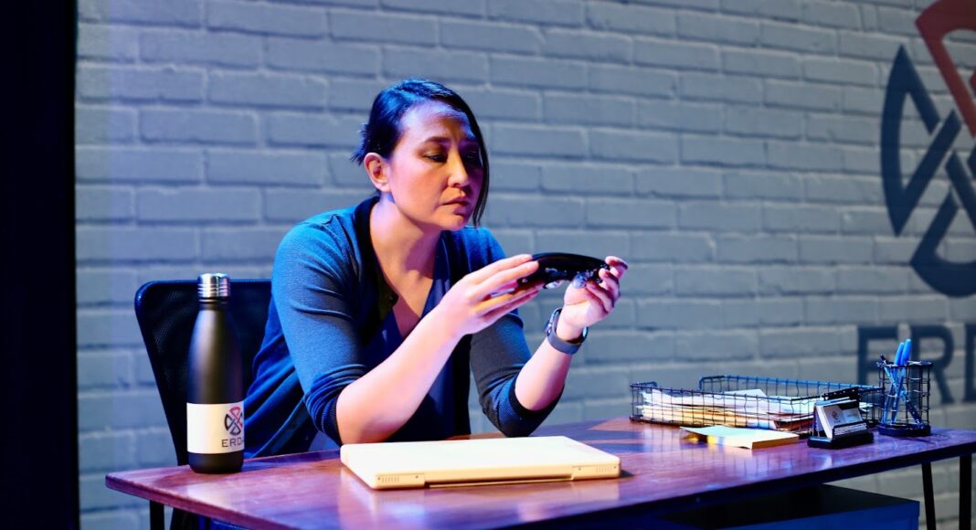 A woman sits at a desk holding a black object, looking at it thoughtfully. The desk has a water bottle, folder, wire basket with papers, a pencil, and other office items. The background has a light brick wall.